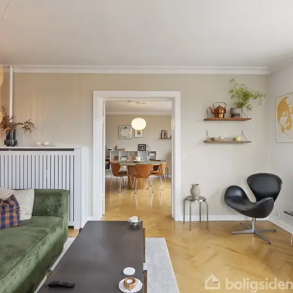 Grøn sofa står i en stue med moderne indretning; der er dekorative lamper over sofaen, åbning fører til spisestue med træbord og stole, og en sort stol i hjørnet ved vinduet.