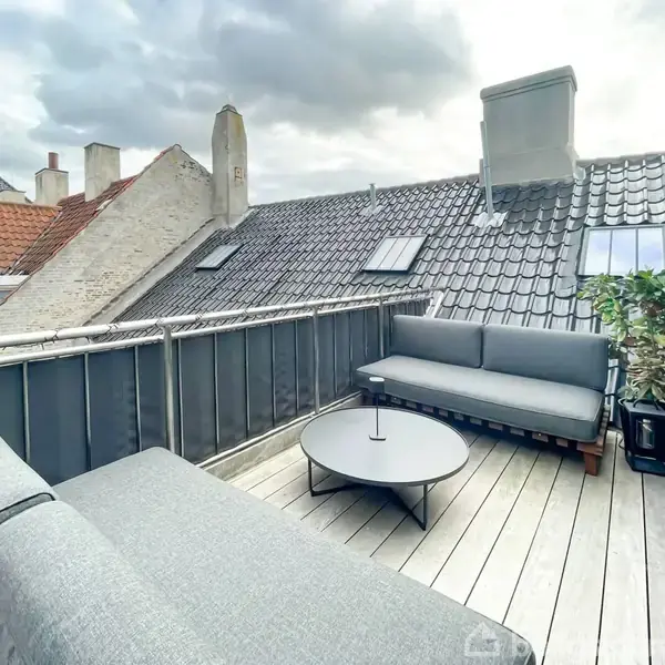 En tagterrasse med grå sofaer omkring et rundt bord, omringet af mørke metalhegn og med udsigt til hustage. Skyet himmel giver en afslappet atmosfære.