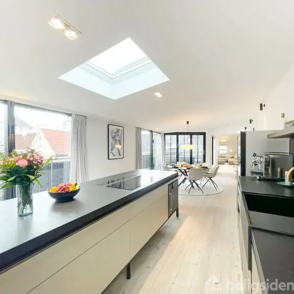 Et moderne køkken med en stor køkkenø og indbyggede apparater; blomster og frugt står på bordet. Rummet er lyst med ovenlysvindue og store vinduer, udsigt til terrasse.