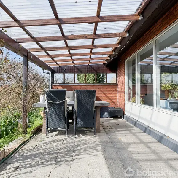 Terrassemøbler står ved et bord under en gennemsigtig overdækning. Terrassefliser ligger i et solrigt område med udsigt til buske og en murstensvæg. Store vinduer reflekterer belysningen.