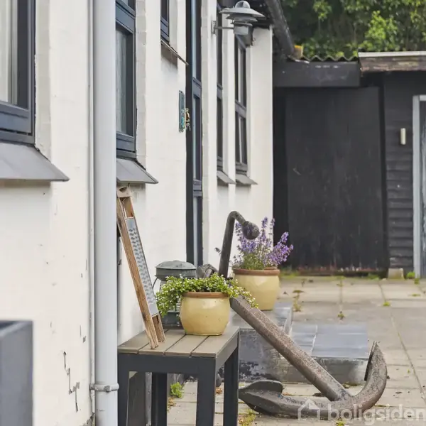 En træbænk med blomsterkrukker og et gammelt anker står langs en hvidmalet bygning med sorte vinduesrammer. Bygningen ligger på en flisebelagt gårdsplads med grønne træer i baggrunden.