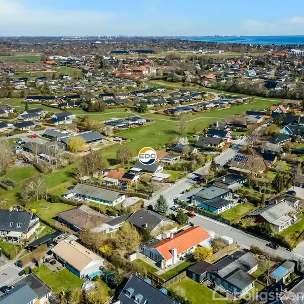 Landskabsudsigt viser et boligområde med spredte huse omgivet af grønne områder og veje. "EDC" logo er placeret over et centralt hus. Byen og havet ses i baggrunden.