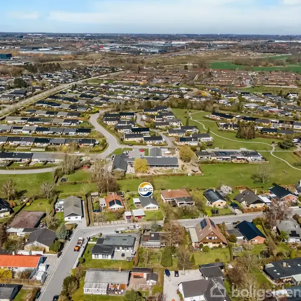 En boligblok ses fra luften, omgivet af en større bebyggelse. Landskabet er grønt med adskillige veje, huse og marker i baggrunden. EDC-skilt er placeret i midten.