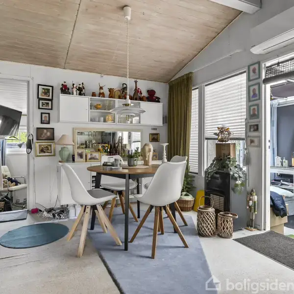 Et rundt træbord med to hvide stole står i en lys stue. Hylder med dekorationer og fotos ved væggen. Brændeovn til højre, dør åbner til overdækket terrasse.