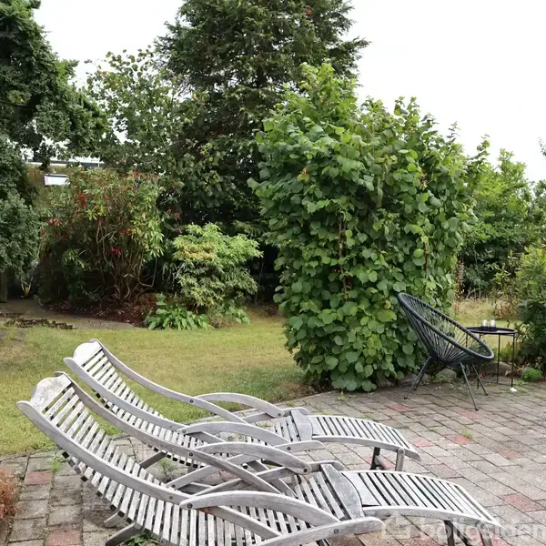 Fire liggestole af træ står på en brolagt terrasse i en have med frodig beplantning. En rund metalstol og bålplads er også synlige.