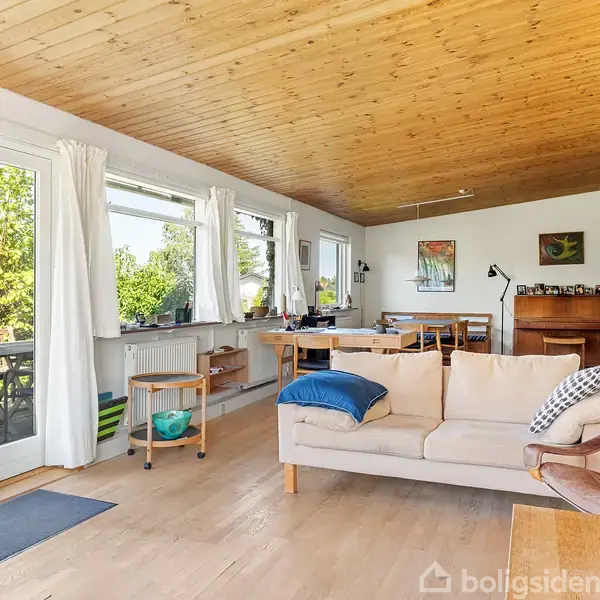 Sofa med puder står foran store vinduer og terrassedøre, som åbner ud mod en grøn have. Rummet har trægulve og -loft, med spisebord i baggrunden.