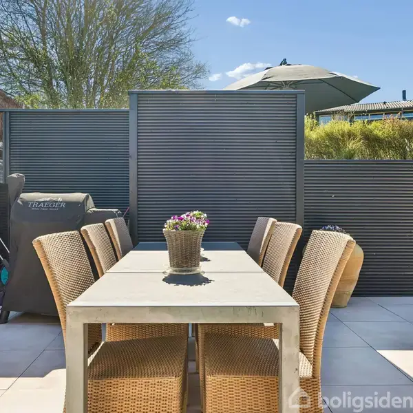 Et udendørs spisebord med seks kurvestole står på en flisebelagt terrasse. En grill med mærket "Traeger" ses til venstre, og der er planter i baggrunden.