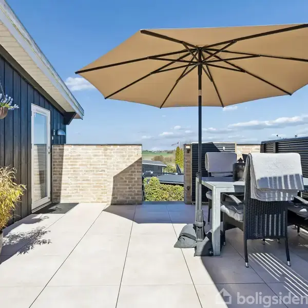 Terrassemøbler står under en stor parasol på en flisebelagt terrasse. En sort træfacade og en murstensvæg danner ramme, mens blomster hænger fra væggen. Solrig himmel ovenover.
