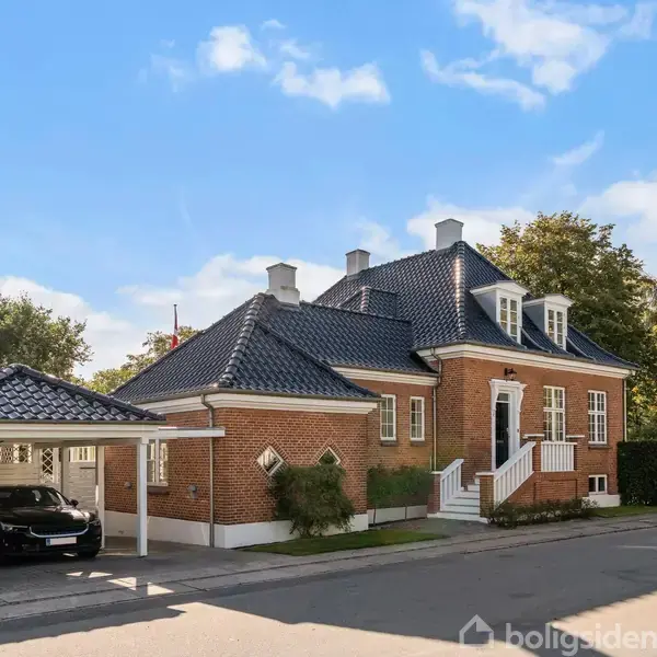 En rød murstensvilla med sort tegltag står i solskin, omgivet af træer. En carport ved siden af huset huser en sort bil.