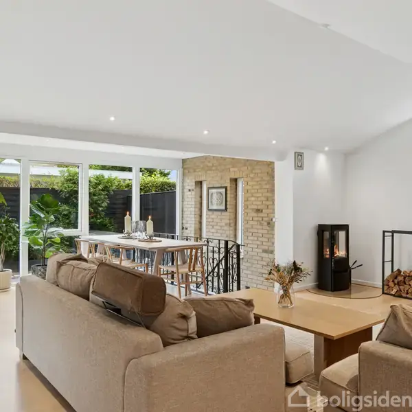 En moderne stue med beige sofaer er arrangeret omkring et sofabord. En brændeovn står ved en murstensvæg, og store vinduer lader lys ind over et bord og planter.