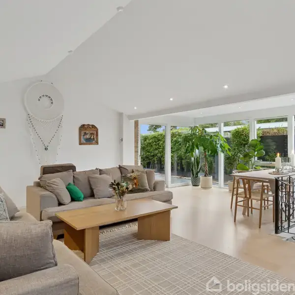 En lys stue indeholder en sofagruppe med farverige puder, et træsofabord med blomster, og en spiseplads ved store vinduer med udsigt til en grøn have.