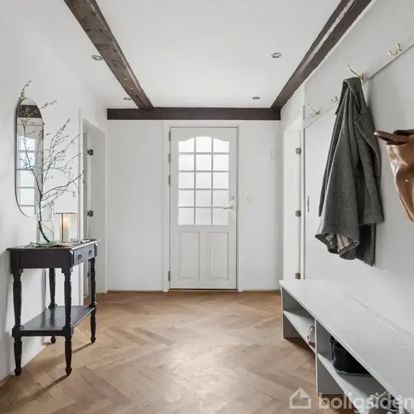 En entré med en hvid dør og glasruder. En bænk med kroge bærer en taske og jakke. Et spejl hænger over et bord med en vase. Gulvet har parketmønster.