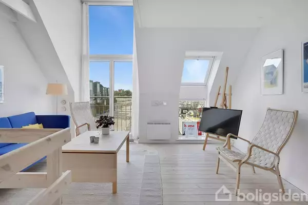 Sofaen står i en lys stue med et stort vindue. To stole og et tv er placeret ved siden af et skråvindue, omgivet af minimalistisk indretning.