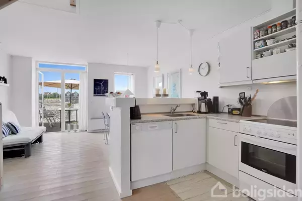 Køkken med hvide skabe og apparater, herunder en ovn og opvaskemaskine; udsigt til stue med spisebord og terrassedøre, der fører ud til altan med paraplyer.