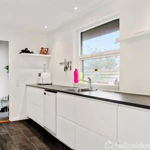 Køkken med hvide skabe og sort bordplade, er udstyret med vask og opvaskemaskine. Lyst interiør med vindue, hylde med små pyntegenstande, og en plantepotte.