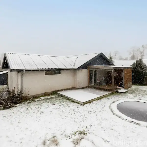 Hus dækket af sne, står stille i en vinterklædt have. Omkringliggende hegn og sparsomme træer i baggrunden skaber en kølig, rolig atmosfære.