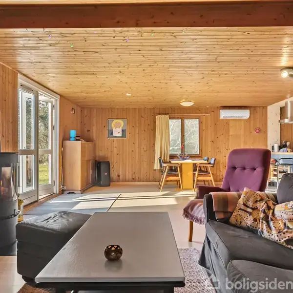 En sofagruppe med bord og stol står i en træbeklædt stue. En brændeovn er tændt ved siden af, mens et spisebord er placeret nær vinduer, der kigger ud til haven.