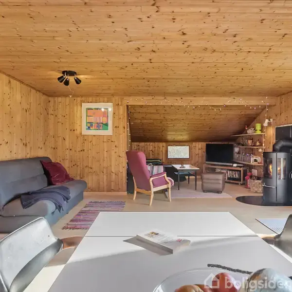 Stue med trævægge og loft, sofa og lænestol mod en brændeovn. Et spisebord i forgrunden er dækket med frugt. Rummet er lyst og indbydende med tæpper på gulvet.