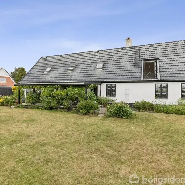 En hvid villa med mørkegråt tag har en overdækket terrasse med grønne planter foran. Den ligger i en have med græsplæne og blomsterbede.