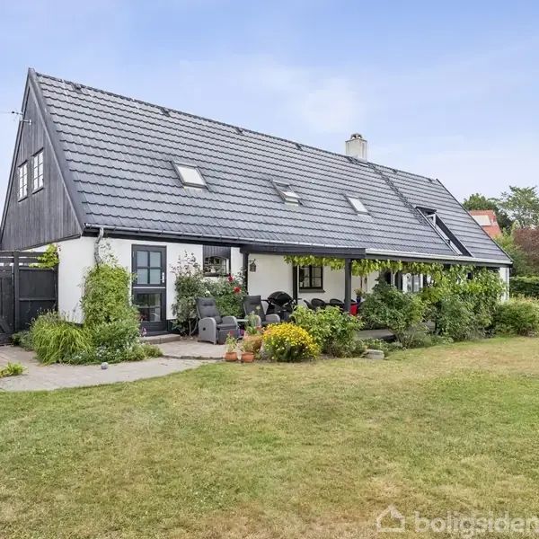 Et lyst, hyggeligt hus med sort tag ligger i en frodig have med græsplæne og buske. Haven har terrasseområde med havemøbler og blomster.