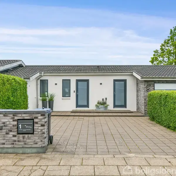 En moderne bungalow har en bred indkørsel af fliser omkranset af velplejede hække. En postkasse ved indkørslen viser nummeret "1". Huset har grå mursten og hvide facader.