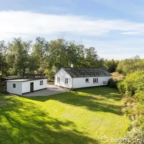 Et hvidt hus og udhus står stille omgivet af grønne træer og en velplejet græsplæne under en klar himmel.
