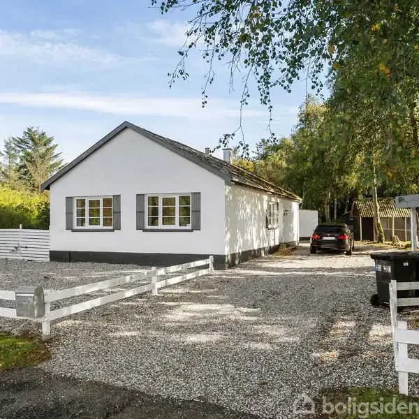 Hvidt hus med grusindkørsel omgivet af træer og grønne buske. Flere biler er parkeret langs indkørslen, hvoraf én er under et halvtag til højre.