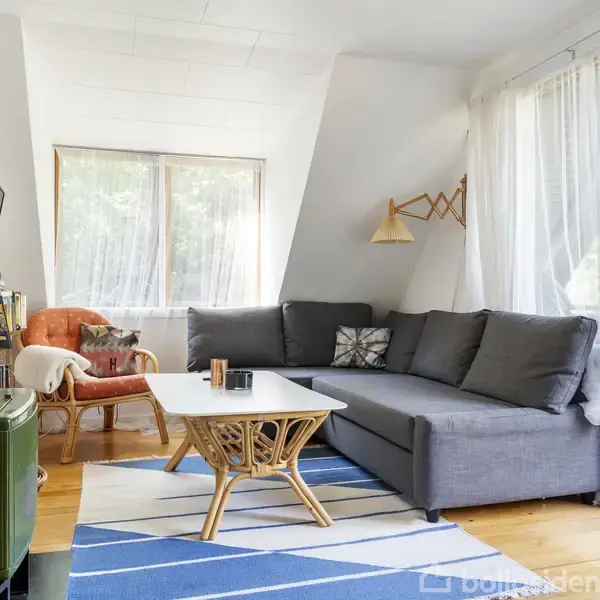 Grønt komfur står i en lys stue med grå hjørnesofa ved siden af et træbord. Store vinduer og åbne døre viser en grøn udsigt, og et tæppe dekorerer gulvet.
