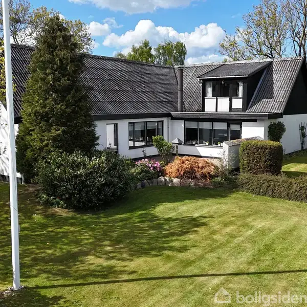 En hvidmalet villa med sort tag ses omgivet af velplejede græsplæner og buske under en blå himmel med lette skyer. Der står et flagstang til venstre for huset.