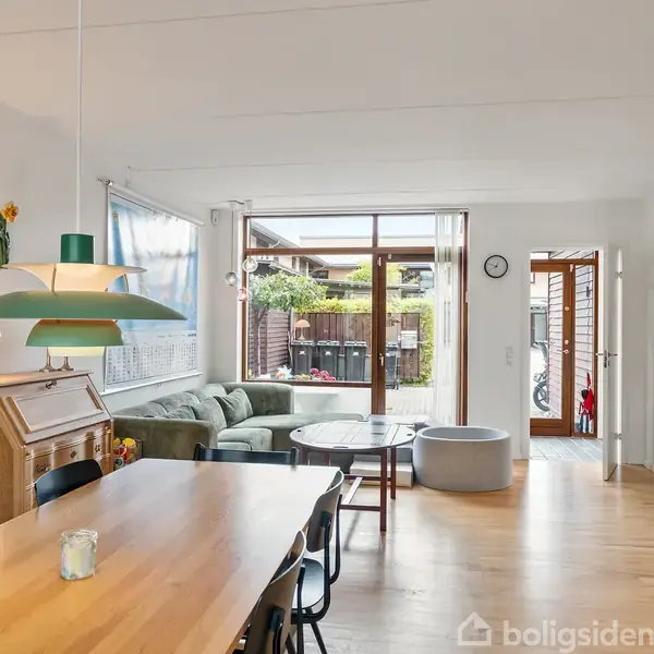 Spisebord med sorte stole står i et lyst stueområde med trægulv og grøn lampe. En sofa og sofabord står ved store vinduer, der fører til en terrasse med planter.