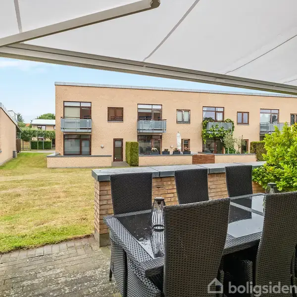 En udendørs spisebordssæt med seks stole står under en markise. Der er en grill til venstre. I baggrunden ses rækkehuse med altaner og en velplejet have.