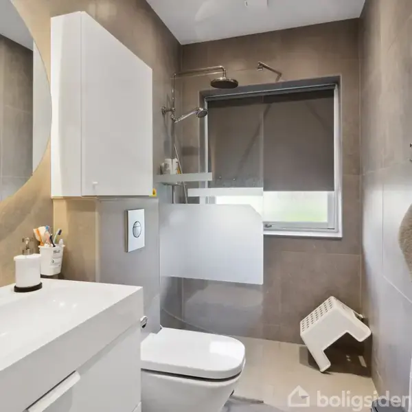 Badeværelse med hvidt skabsarrangement, stort rundt spejl og håndvask. Et toilet er ved siden af. Et vindue med rullegardin giver lys, og en brusekabine er i hjørnet.