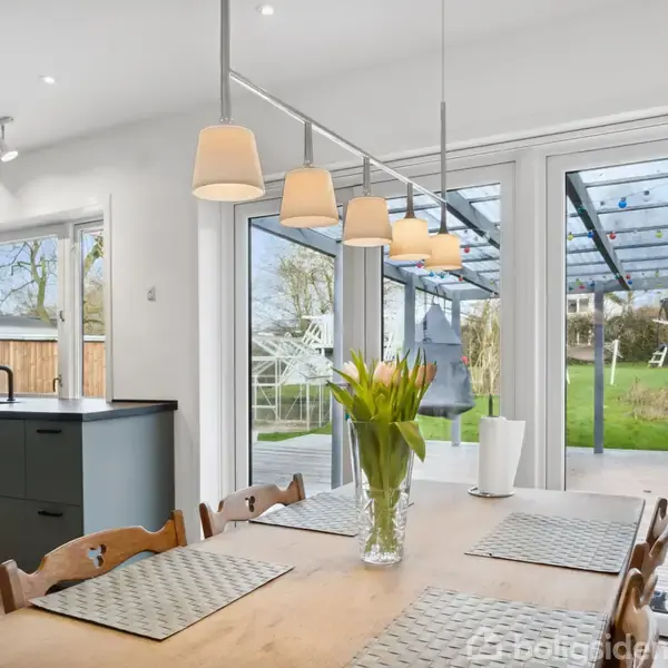 Et lyst køkken-alrum med træbord og blomster i centrum. Store glasdøre åbner mod en have med græsplæne og overdækket terrasse. Moderne køkkenskabe og hængende lamper fuldender rummet.