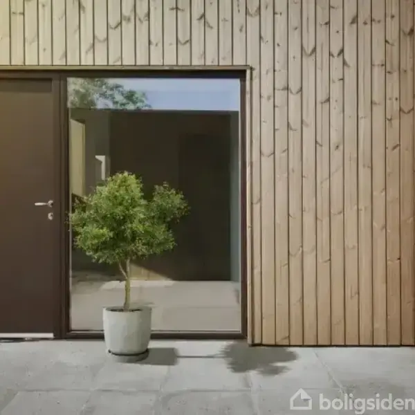 En dør og et stort vindue dominerer en træbeklædt facade. En grøn plante i en hvid potte står foran døren på en betonterrasse.