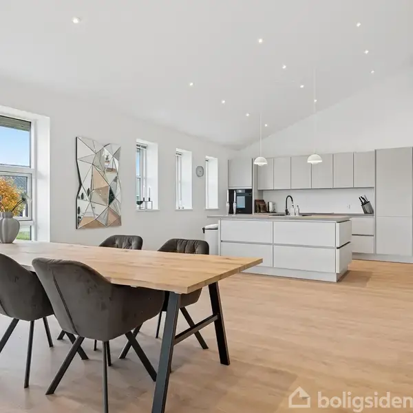 Spisebord med fire stole står i en lys, moderne stue-køkken. Store vinduer viser en have med træer. Et geometrisk spejl hænger på væggen overfor et sofistikeret, minimalistisk køkken.