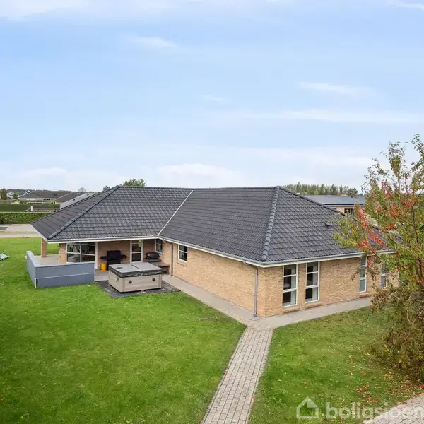 En moderne murstensvilla med gråt tag står på en velplejet græsplæne, omgivet af nogle træer og udendørs møbler. Villaen ligger i et fredeligt boligkvarter.