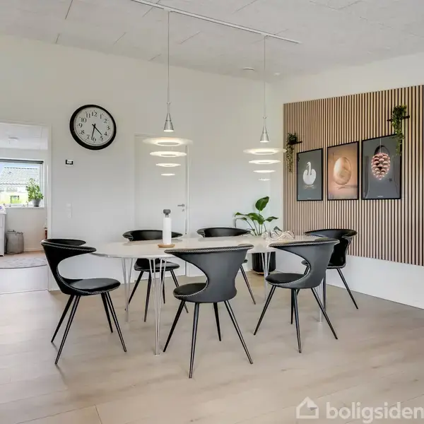 Et rundt bord med sorte stole står i en moderne spisestue. Over bordet hænger tre lamper. Væggene er dekoreret med billeder og en ur. Rummet er lyst og rummeligt.