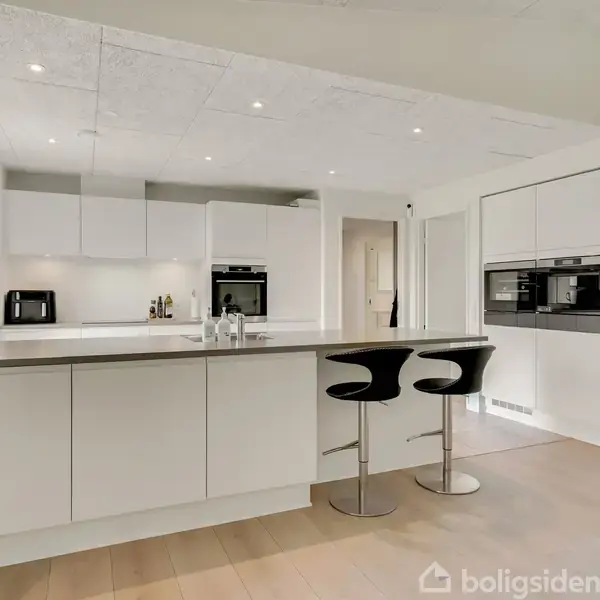 Et moderne køkken med en stor hvid ø, hvorpå der står glas og en vandflaske. To sorte barstole står ved øen. I baggrunden ses skabe og indbyggede ovne.