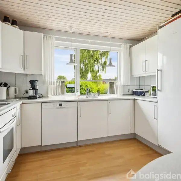 Hvidt køkken med hjørnebordplade og moderne apparater, inklusive ovn og vask. Store vinduer tillader naturligt lys, og en lysekrone hænger over bordet. Udsigt til træer udenfor.