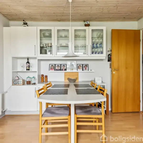 Et spisebord med syv stole står centralt i et spisekøkken. Køkkenskabe med glaslåger og porcelæn hænger på væggen. En åben dør fører til næste rum.