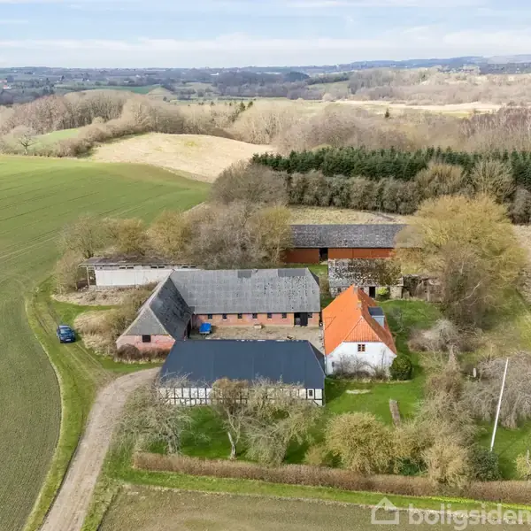 Gårdkompleks beliggende i naturskønne omgivelser med marker og skov. Flere bygninger med røde tage og staldlignende strukturer omgives af træer og grusvej. En bil holder parkeret nær bygningerne.