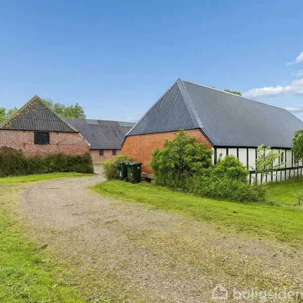 To røde murstensbygninger med sorte tage står ved siden af en grussti omgivet af grønt græs og buske under en klar blå himmel.