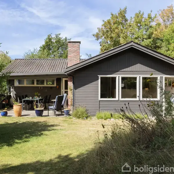 Et træhus står i en have med græsplæne og buske; huset har en terrasse med stole og krukker. Sollys belyser facaden, omgivet af grønne træer.
