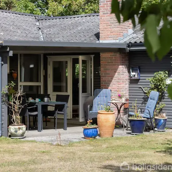 En terrasse med havemøbler og blomsterkrukker foran et moderne mørkt træhus med store vinduer. Solskin omgiver området, der er bevokset med grønne træer og buske.