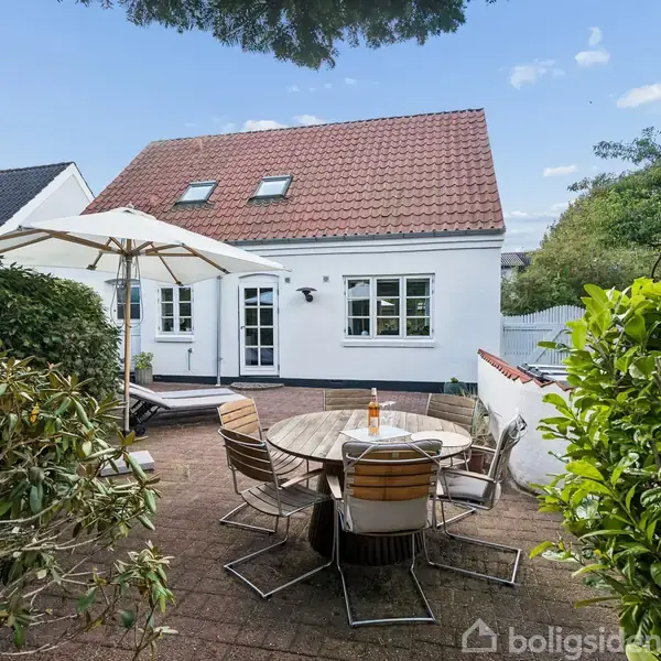 Rundt havebord med stole står på terrasse. Parasol er opslået. Baggrunden viser et hvidt hus med rødt tag. Omgivet af grønne buske under blå himmel.