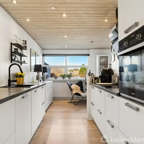 En moderne stue med læderstole omkring et rundt bord, overstrålet af en pendellampe. Store vinduer giver udsigt til terrasse og have. Farverige malerier pynter de hvide vægge.