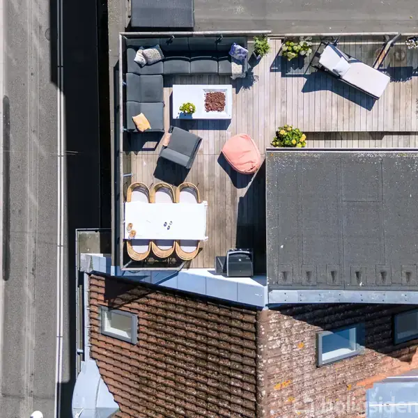En hyggelig tagterrasse med sofaer og borde er indrettet til afslapning omkranset af bybygninger. En person cykler langs gaden ved siden af.