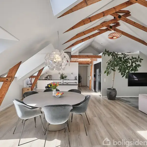 Spisebord med polstrede stole omgivet af en moderne stue med loftsbjælker. Et stort, grønt plante træ pryder rummet, der indeholder et fjernsyn og sofa. Lyse trægulve og spisebordsdekorationer i form af frugt.