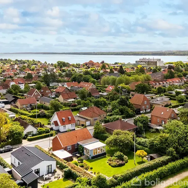 Boligområde med huse med røde tage spredt over grønne haver og træer. I baggrunden anes en sø eller fjord, mens skyer dækker himlen.