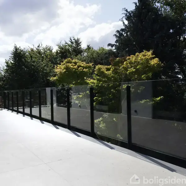 En terrasse med glasrækværk strækker sig langs en frodig have med tætte træer og buske, mens to stole står i venstre hjørne under en delvist skyet himmel.